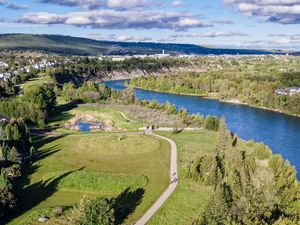 Aerial view of a lush green park along a winding river with tree-lined banks and a city skyline in the background.