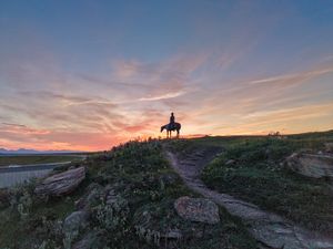 Silhouette of a horse and rider on a rocky hill at sunset with vibrant orange and pink skies over a vast landscape.