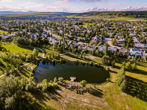 Aerial view of a suburban neighborhood with a serene pond, lush greenery, and mountain backdrop under a partly cloudy sky.