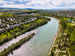 Aerial view of a winding turquoise river flanked by lush green trees and suburban neighborhoods under a partly cloudy sky.