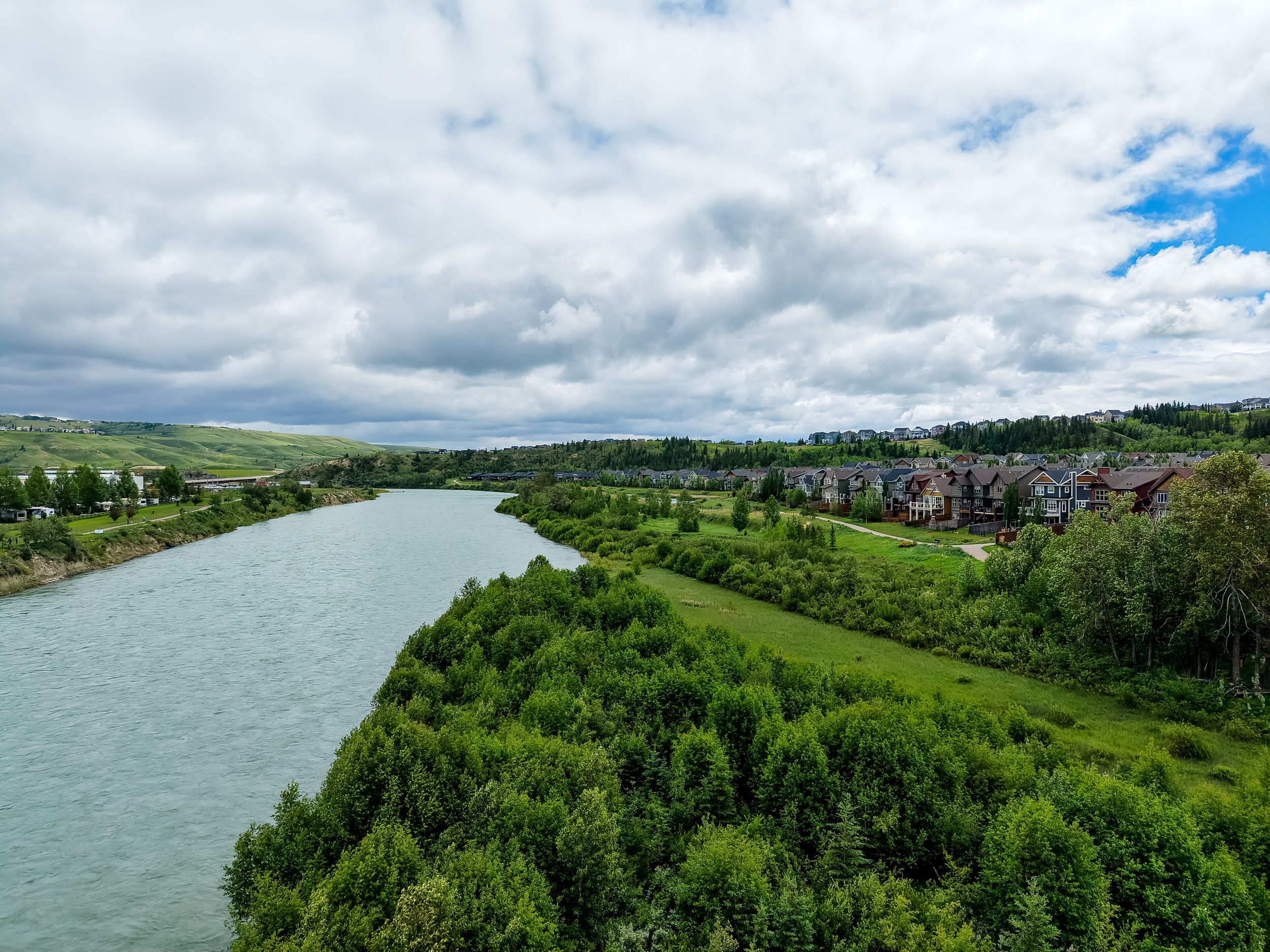 Aerial view of a winding river flanked by lush green trees and a residential neighborhood under a partly cloudy sky.
