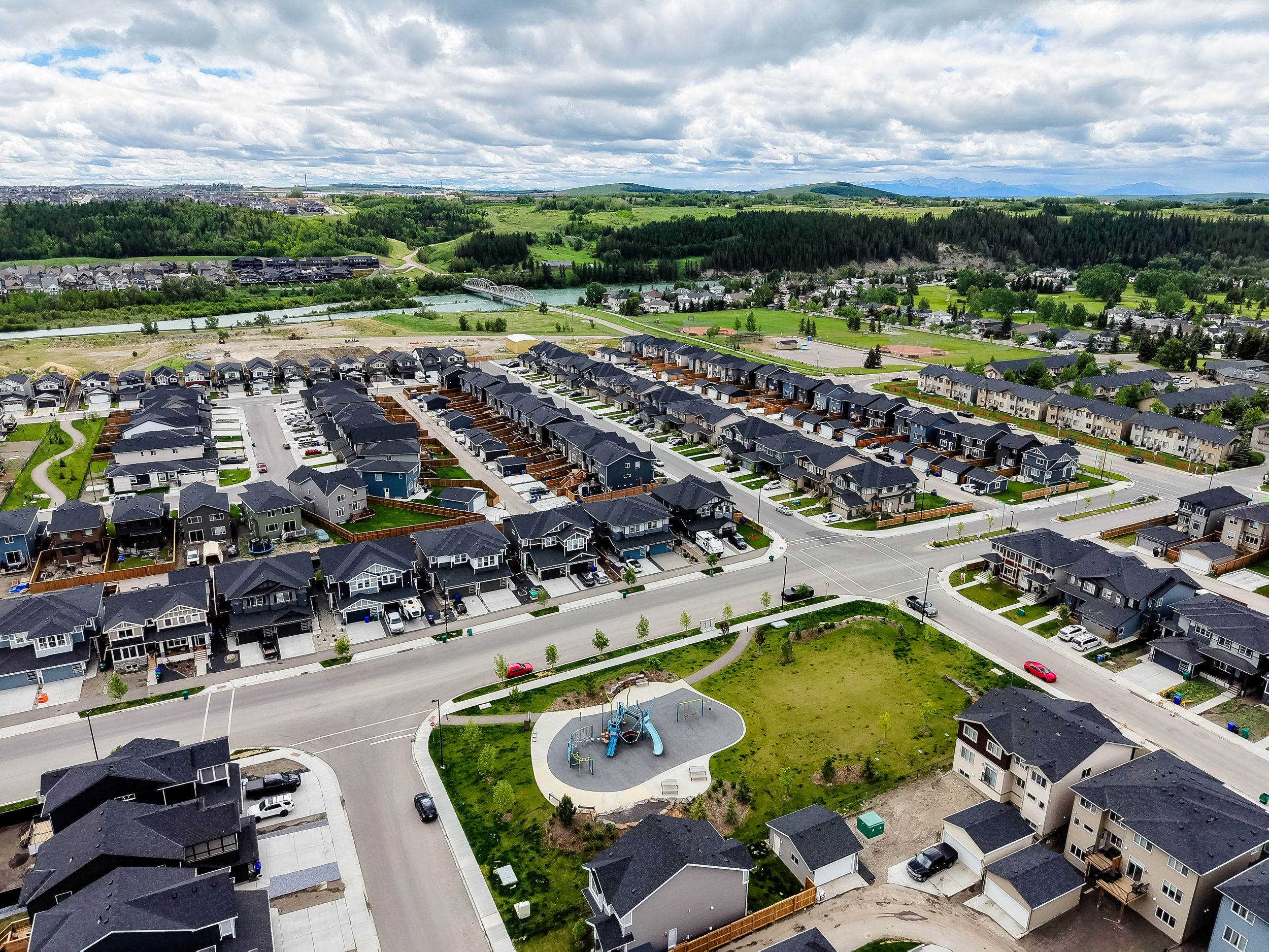 Aerial view of a modern suburban neighborhood with dark-roofed homes, a central fountain plaza, and green hills in the background.
