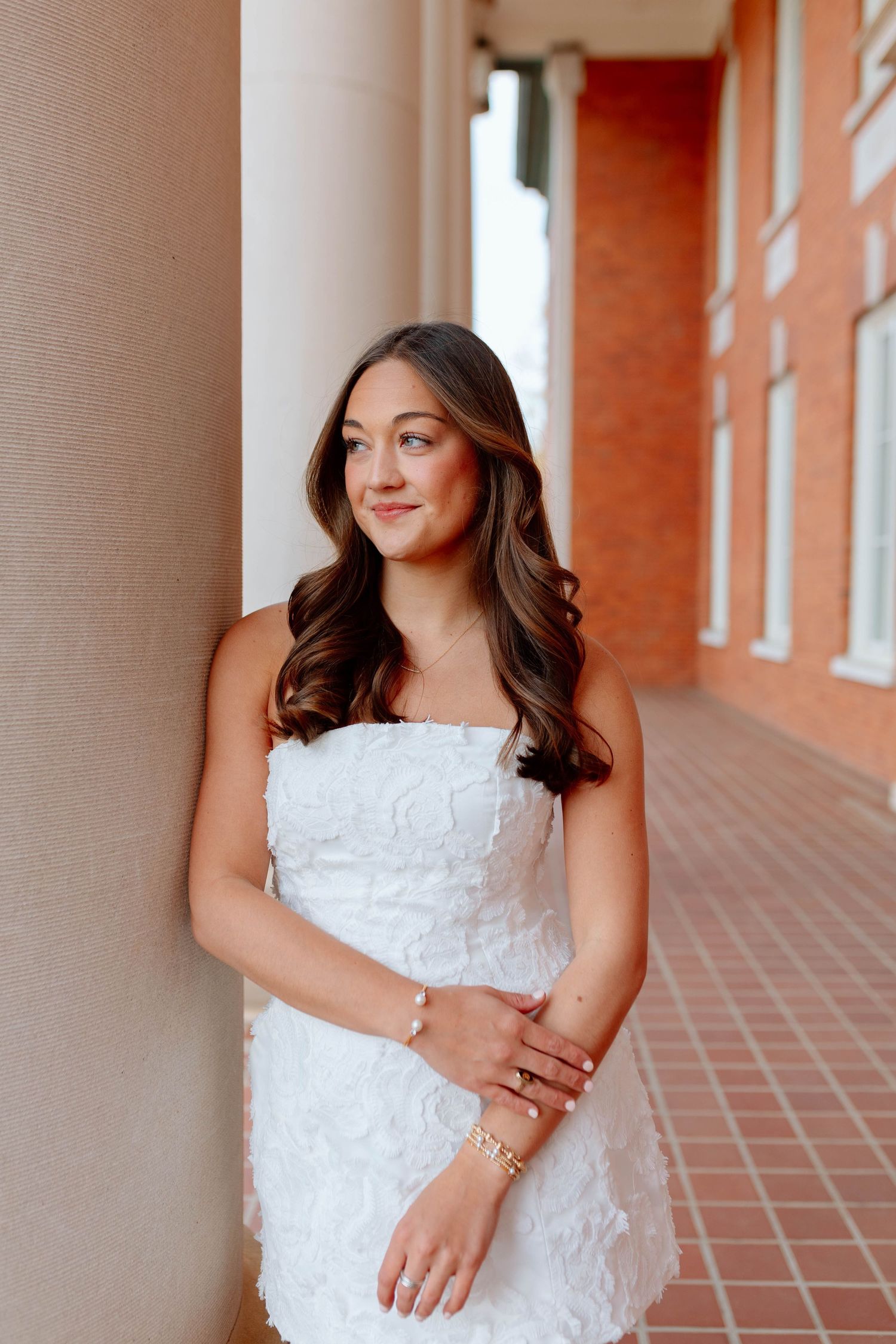 Young woman in white strapless floral dress poses gracefully between columns, wearing gold jewelry, with long wavy hair.