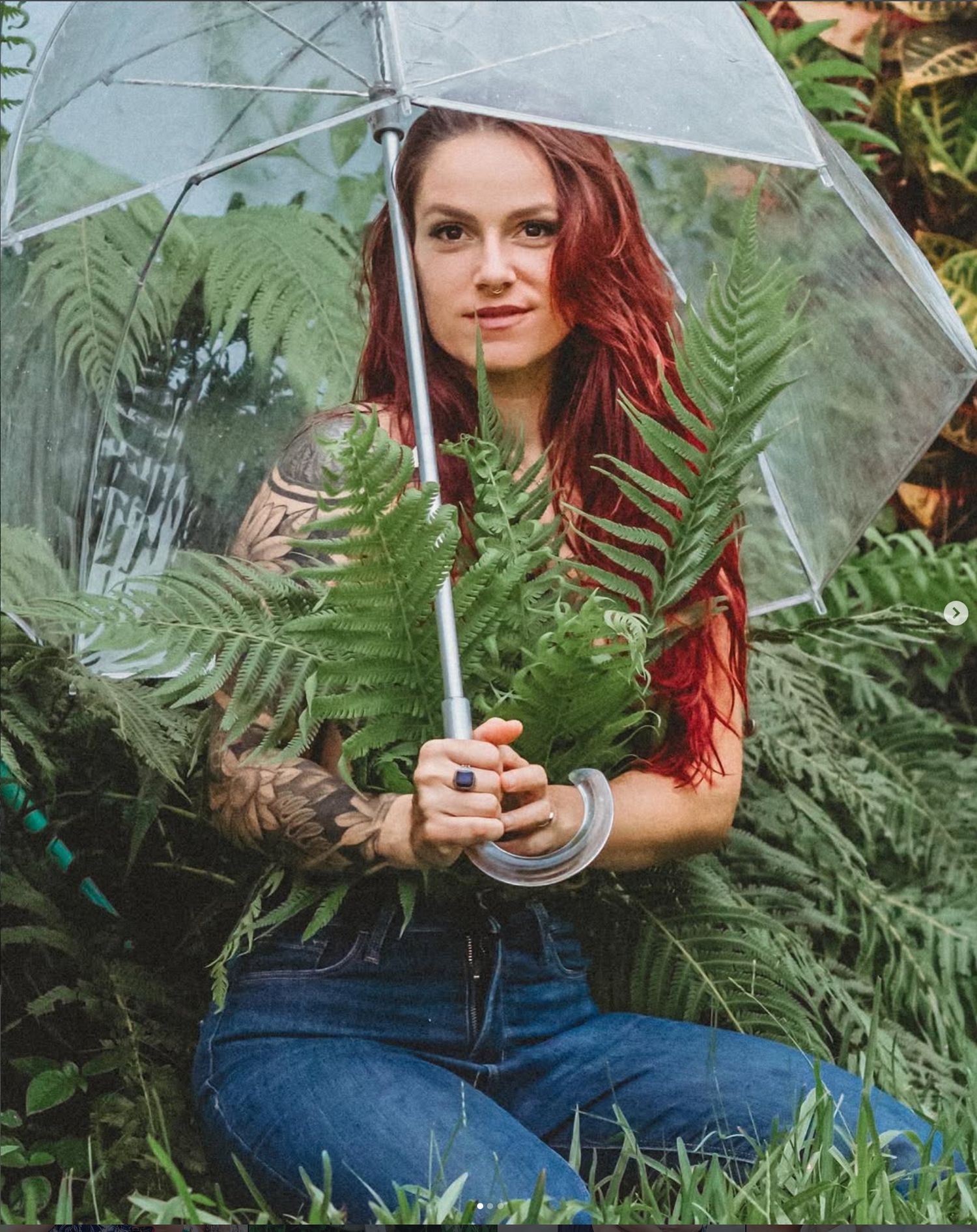 Woman with red hair holds clear umbrella among lush ferns, showcasing tattoos and blue ring outdoors.