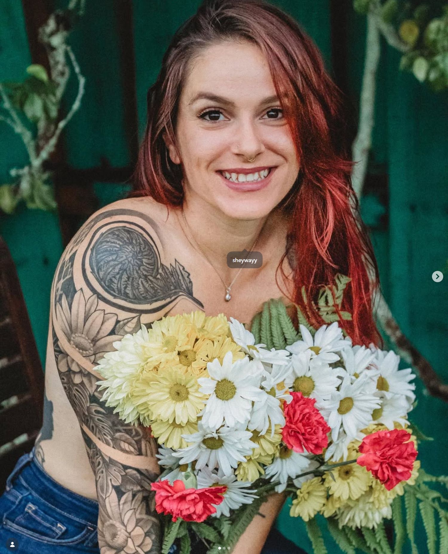 Smiling woman with red hair and floral sleeve tattoo holding a bouquet of daisies and carnations outdoors.
