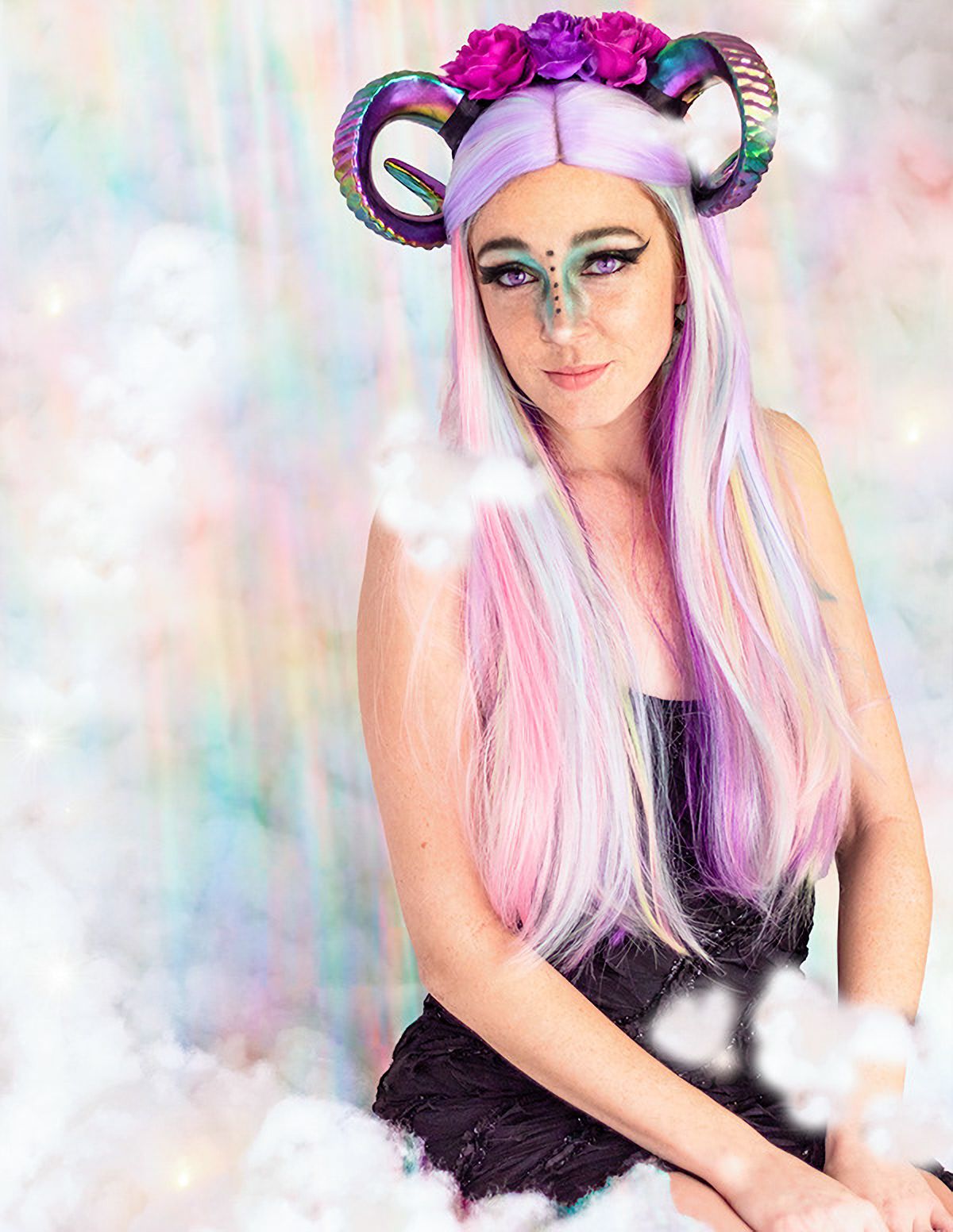 Woman with long pastel pink and purple hair wearing colorful ram horns headpiece and pink flowers against rainbow background.
