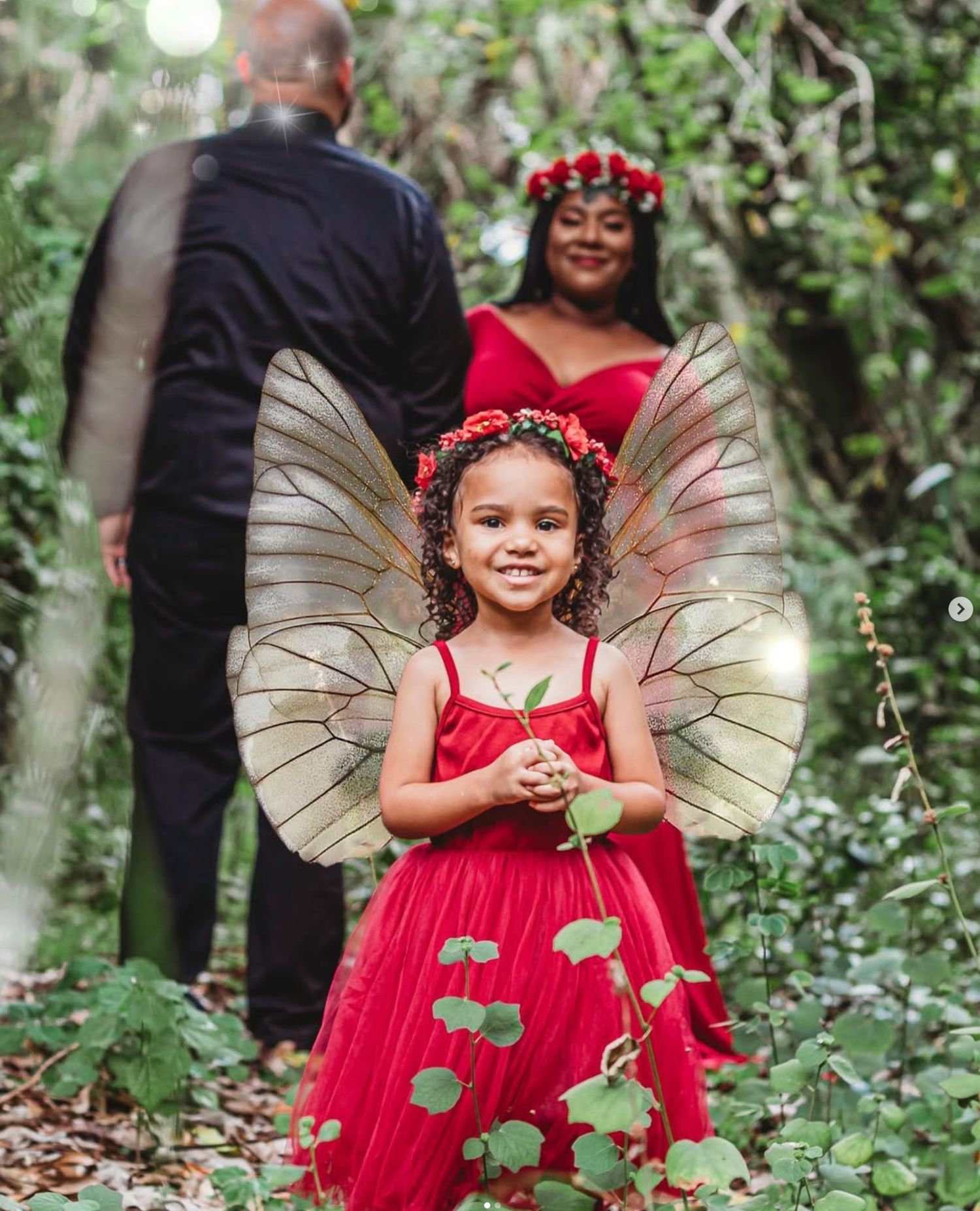 A young girl in a red dress and fairy wings with a floral crown poses in a lush garden with a couple behind her in Riverbend Park, Jupiter.