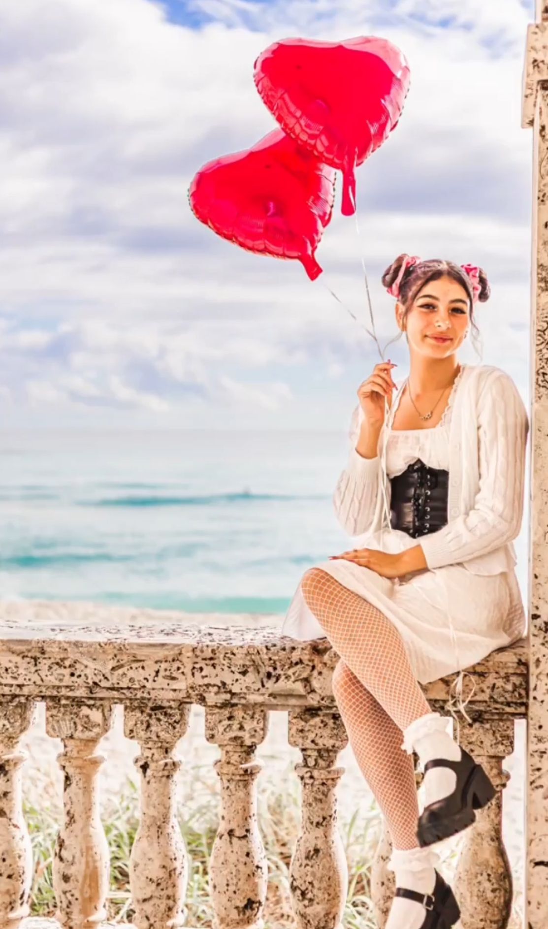 Young woman in white dress and black corset holds red heart balloons while sitting on a stone balustrade by the sea on worth ave palm beach.