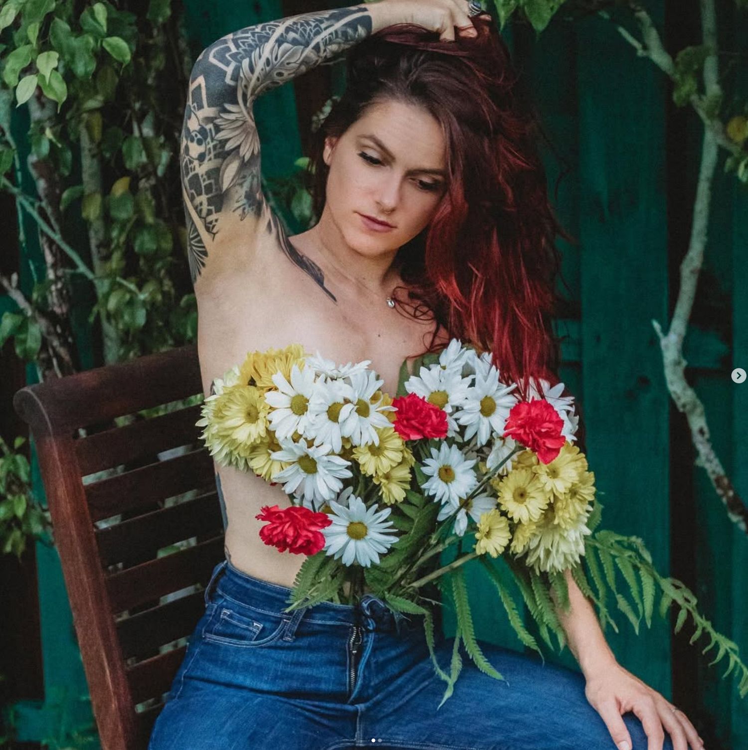 Woman with tattoos and red hair holding colorful daisies and carnations, seated on a wooden chair outdoors.