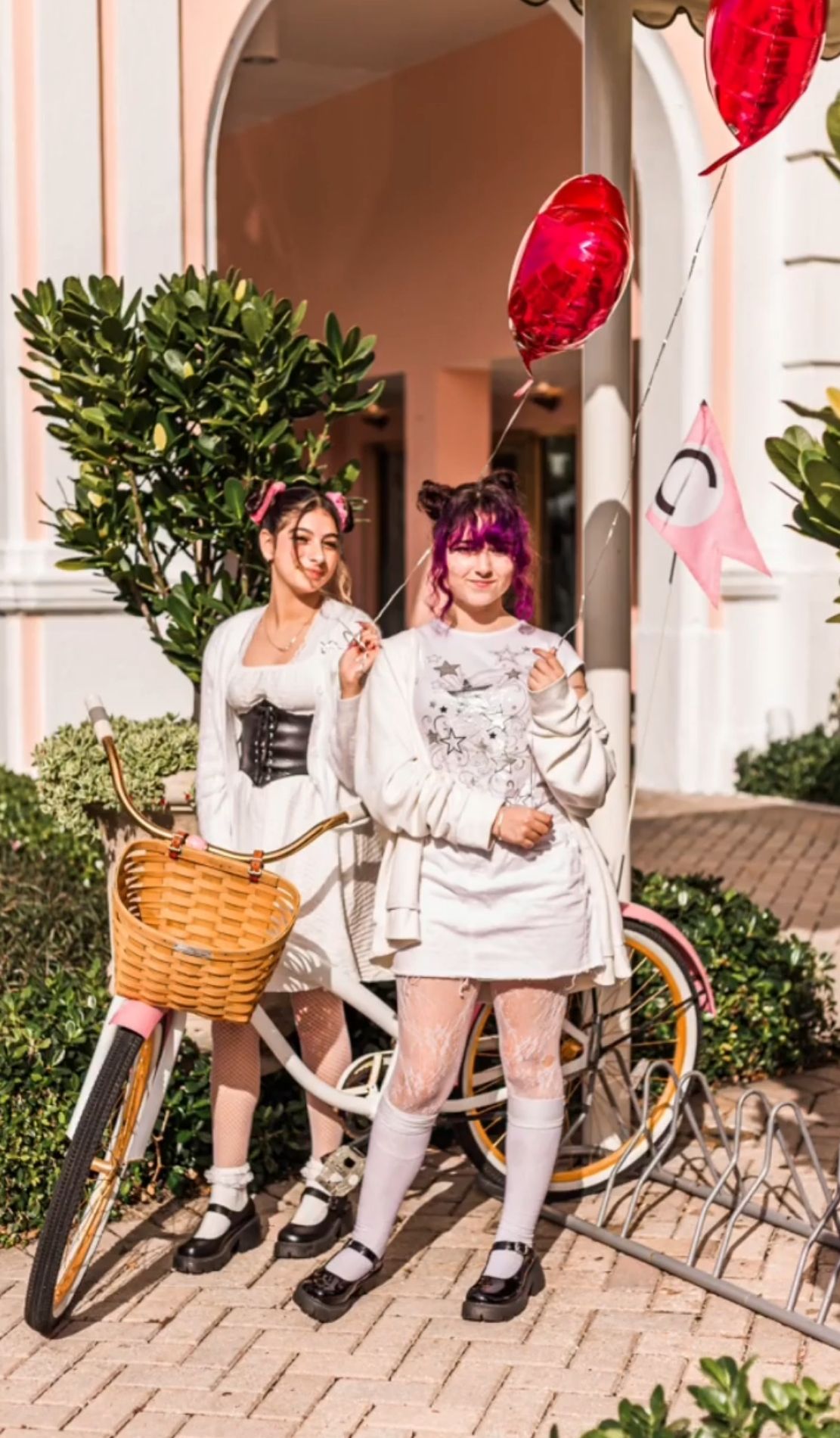 Two young women in white outfits with black corsets pose beside a bicycle with balloons outdoors on worth ave palm beach fl.