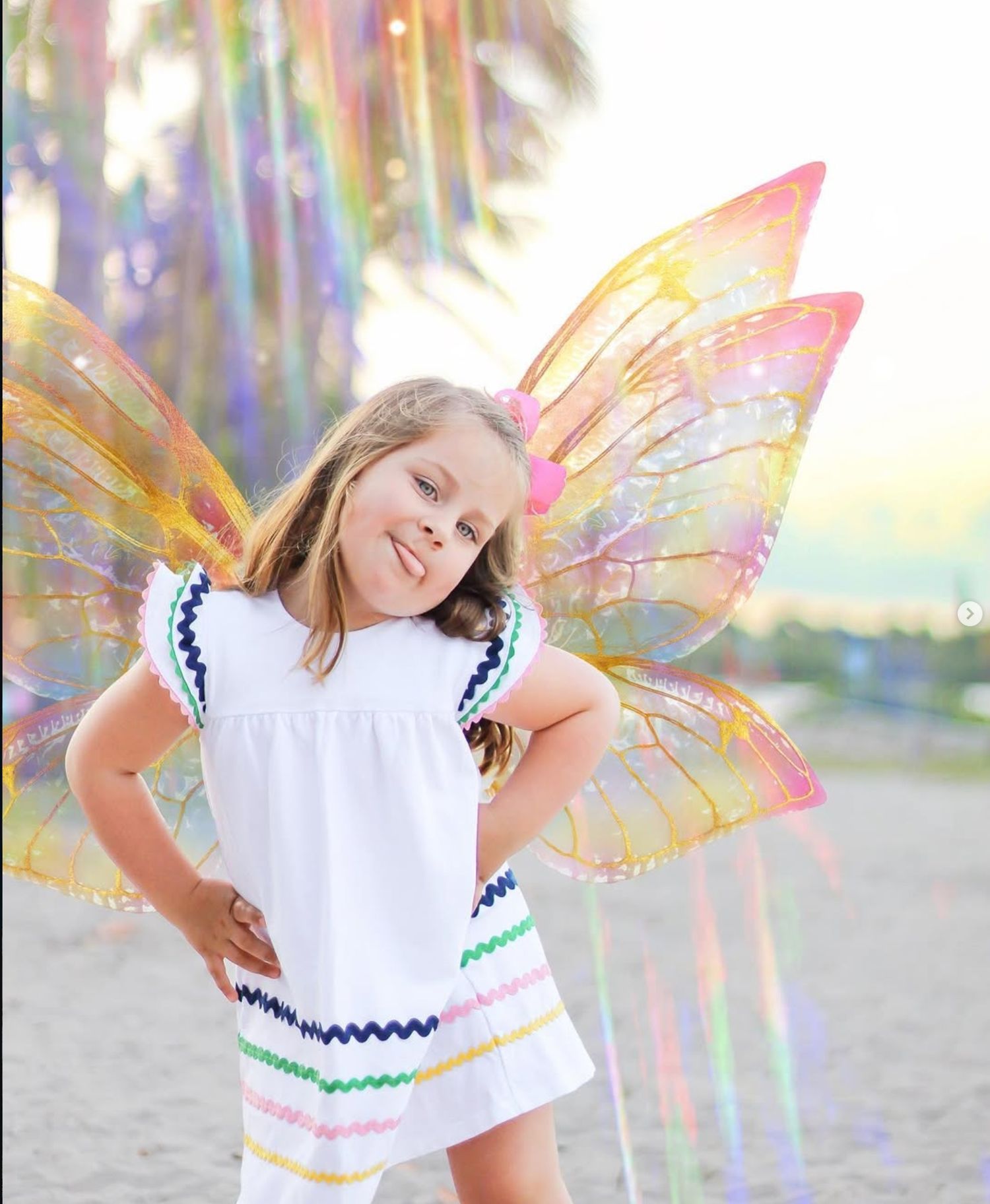 Young girl in white dress with colorful trim poses on Dubois beach wearing iridescent rainbow fairy wings.