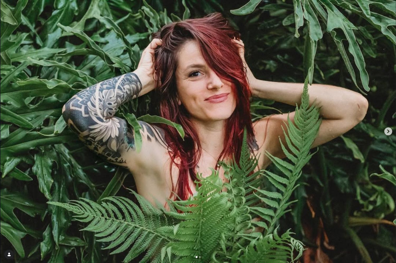 Woman with red hair and tattoos posing among lush green ferns and tropical foliage.