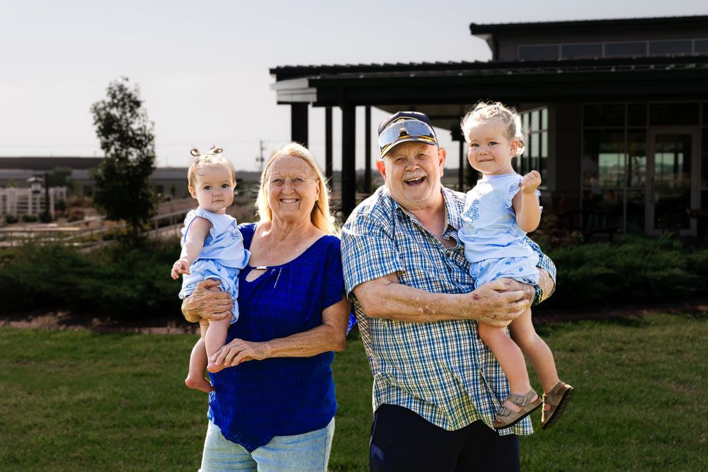 Grandparents smiling outdoors holding two young toddlers in blue outfits with a modern building in the background.