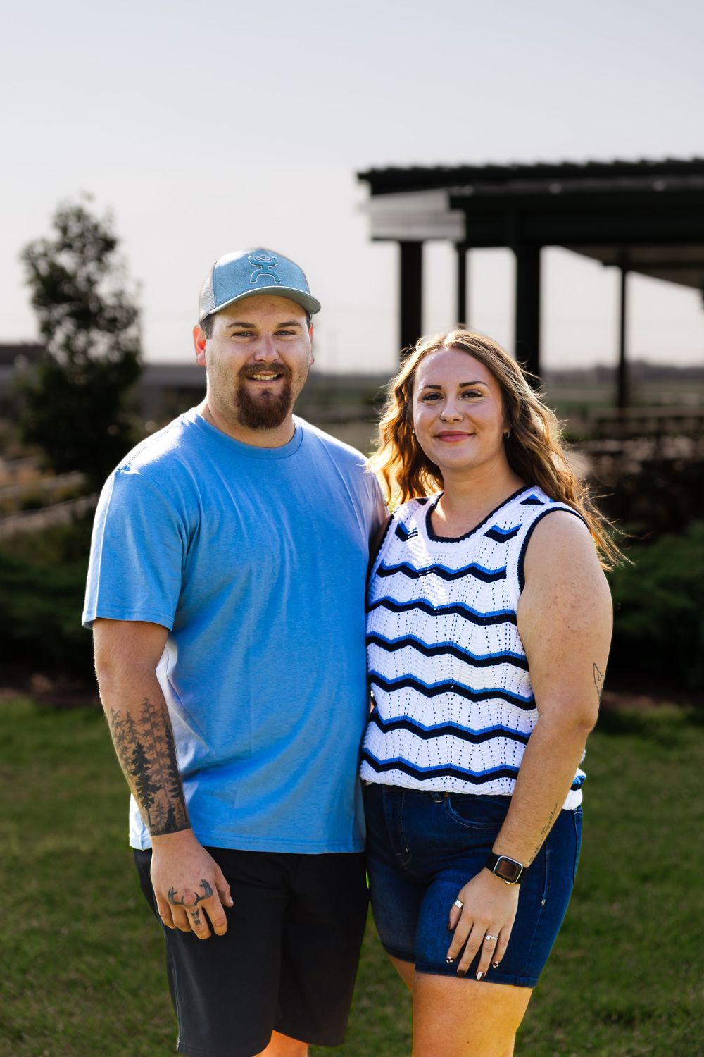 Smiling couple posing outdoors on green grass, man in blue t-shirt and cap, woman in striped top and denim shorts.