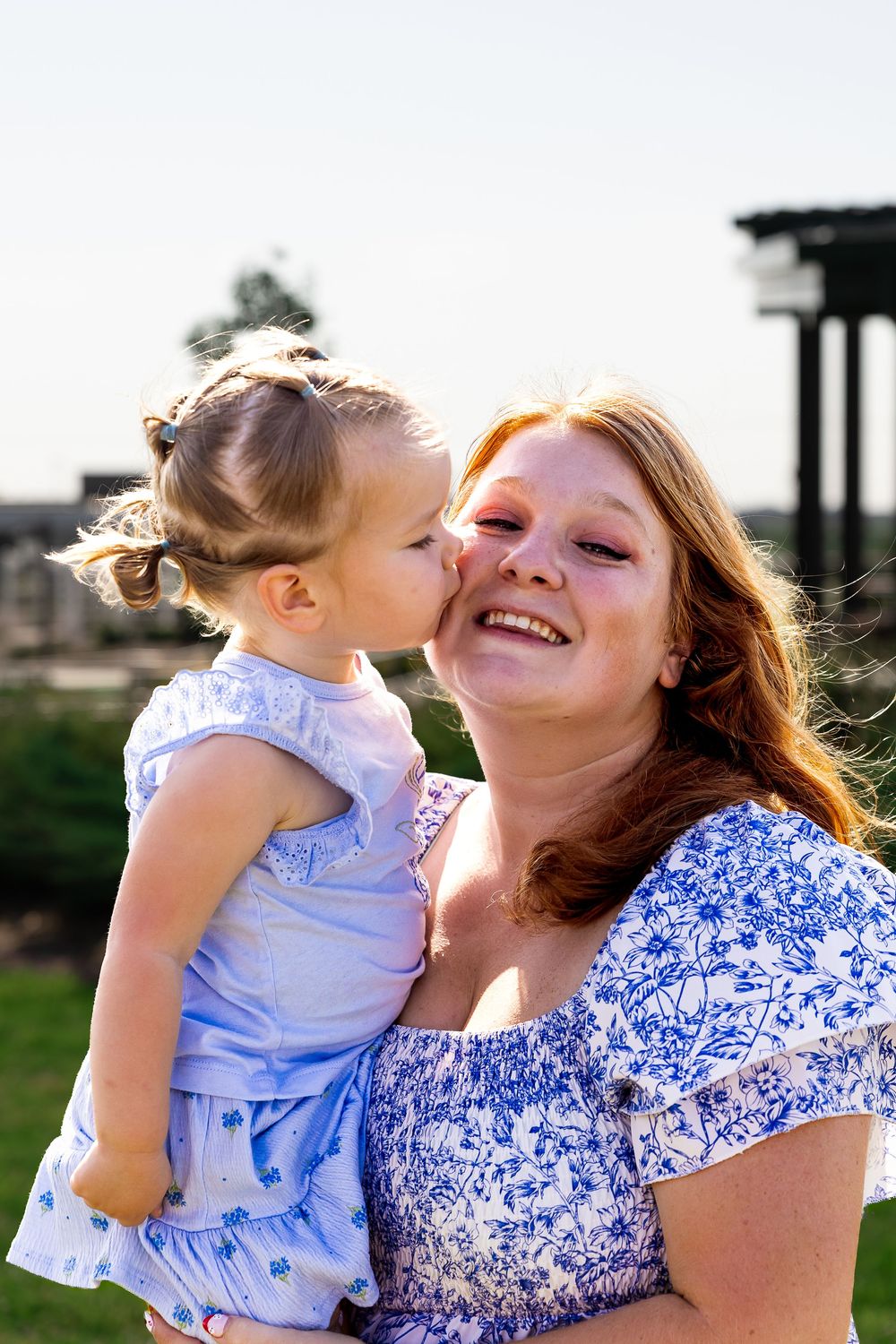 A red-haired woman smiles joyfully while holding a young girl in a blue dress who kisses her cheek outdoors.