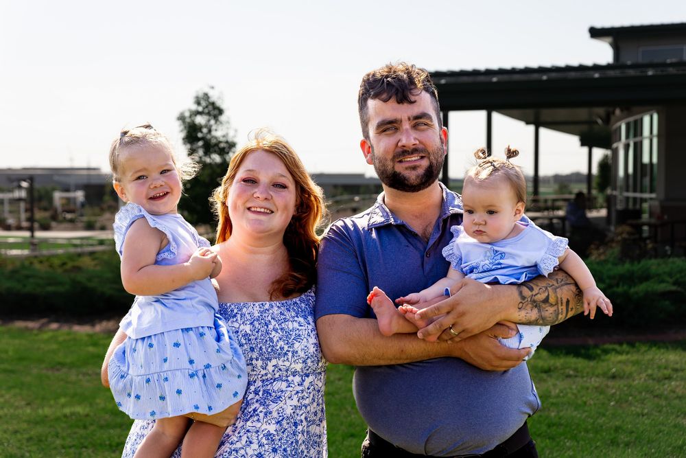 A happy young family of four poses outdoors, with two parents holding their young daughters dressed in blue.