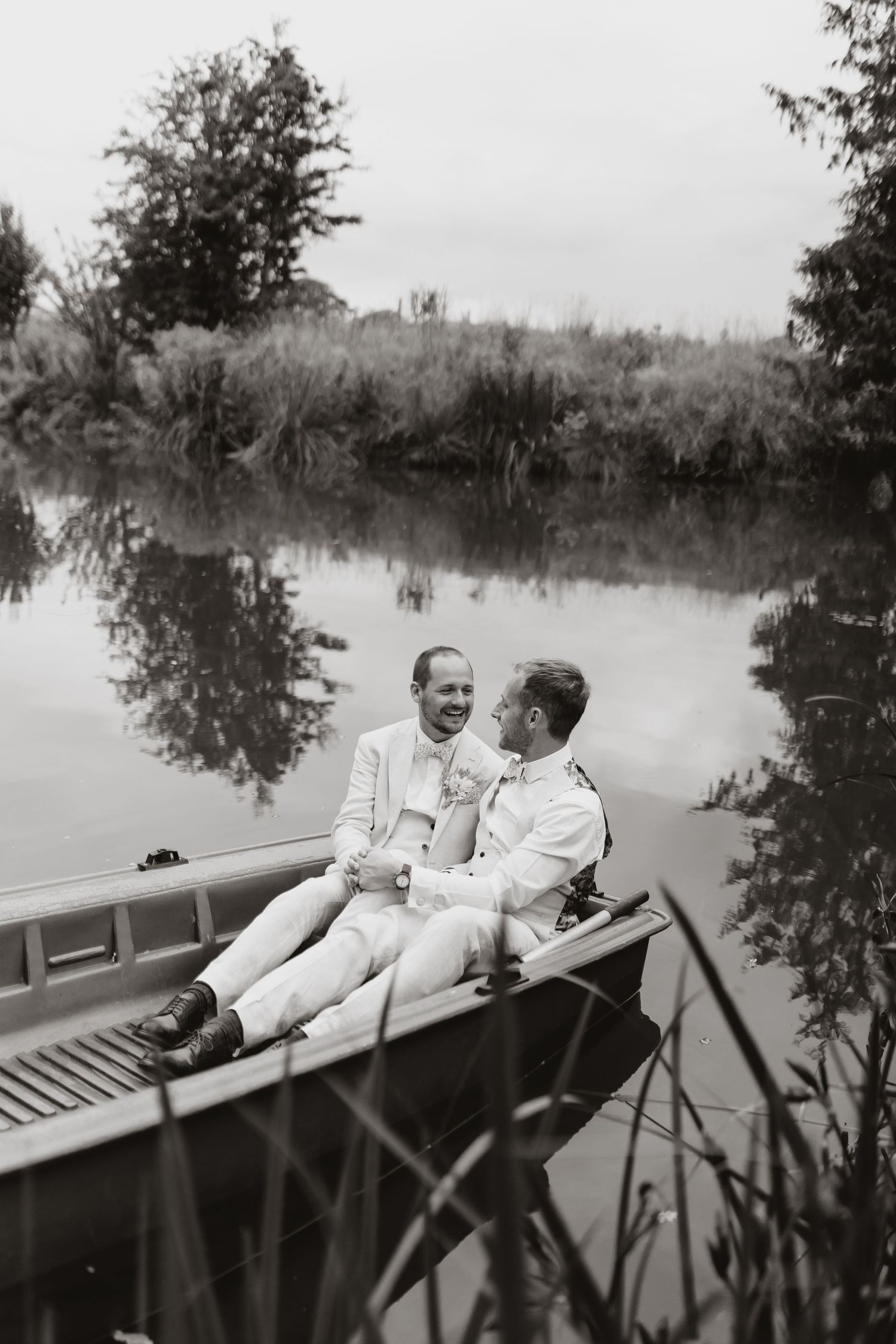 Two grooms laugh together on a boat on a calm river, surrounded by lush greenery, in a black and white wedding photo at Moat Hall Barns