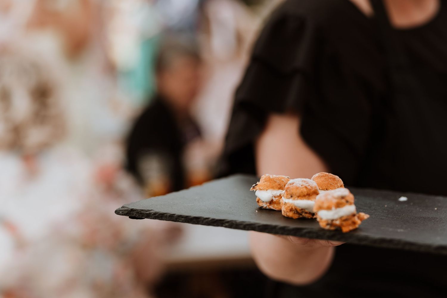 A server in black attire holds a slate board with mini cream-filled sliders at an elegant catering event.