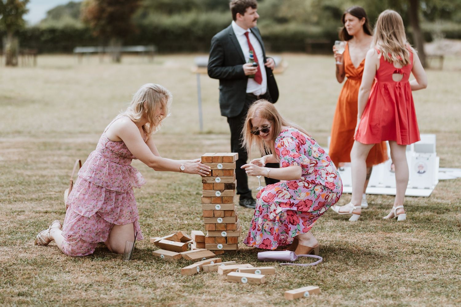 Two women playing giant Jenga at an outdoor wedding reception on a sunny day with guests socializing in background.
