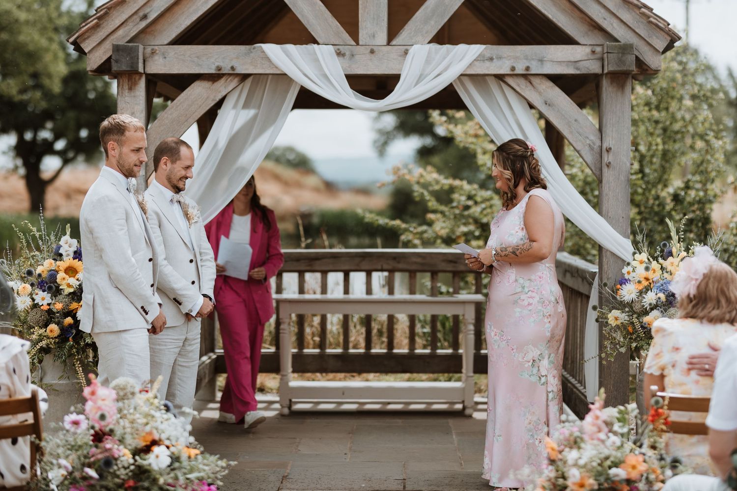 Two grooms in white suits listening to sister say a poem under a rustic wooden arch adorned with flowers and white draping.