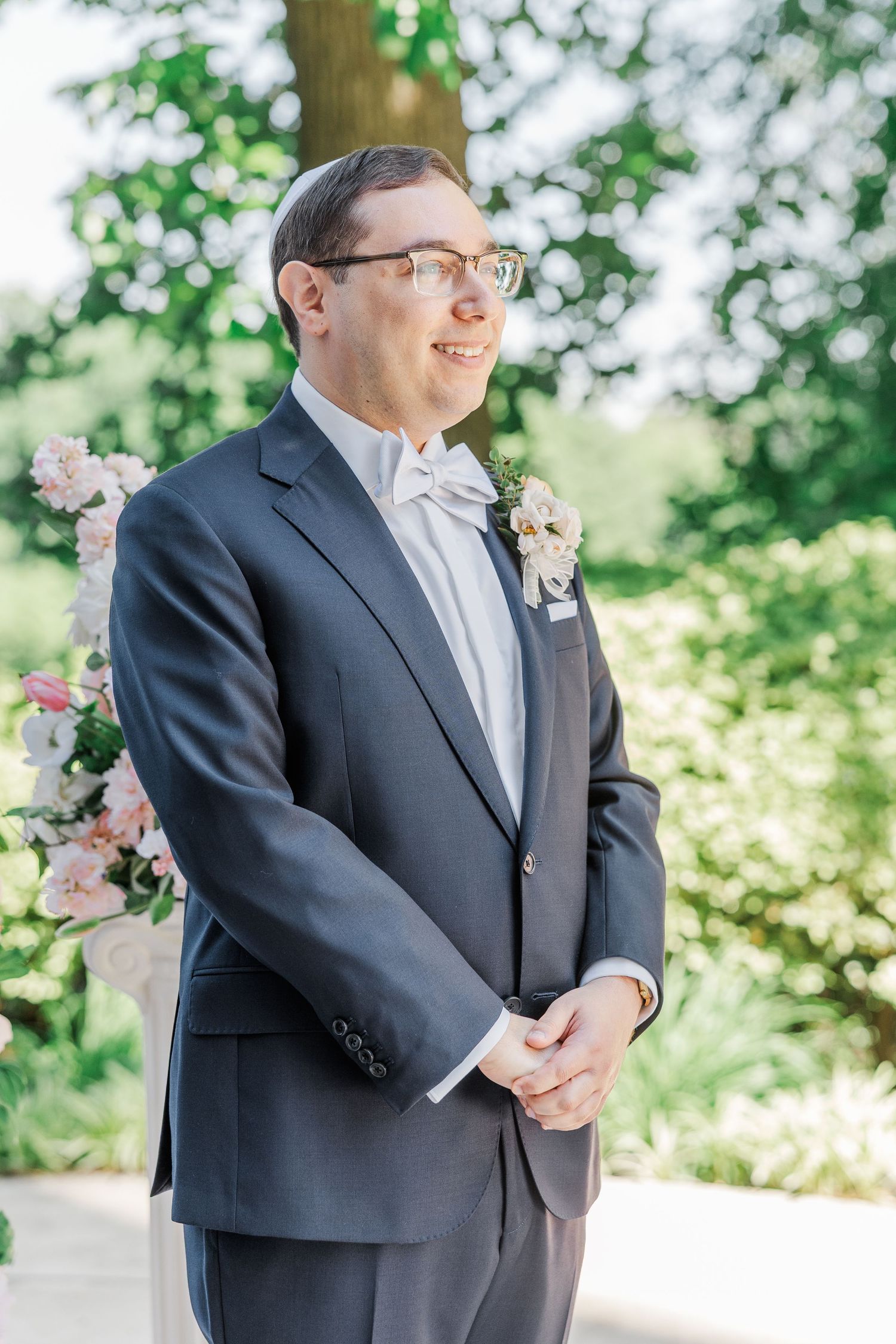 Groom in navy tuxedo with white bow tie and floral boutonniere smiling outdoors at garden wedding ceremony.