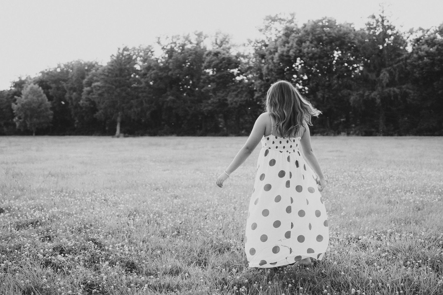 A young girl in a polka dot dress walks through a lush green field with trees in the background.