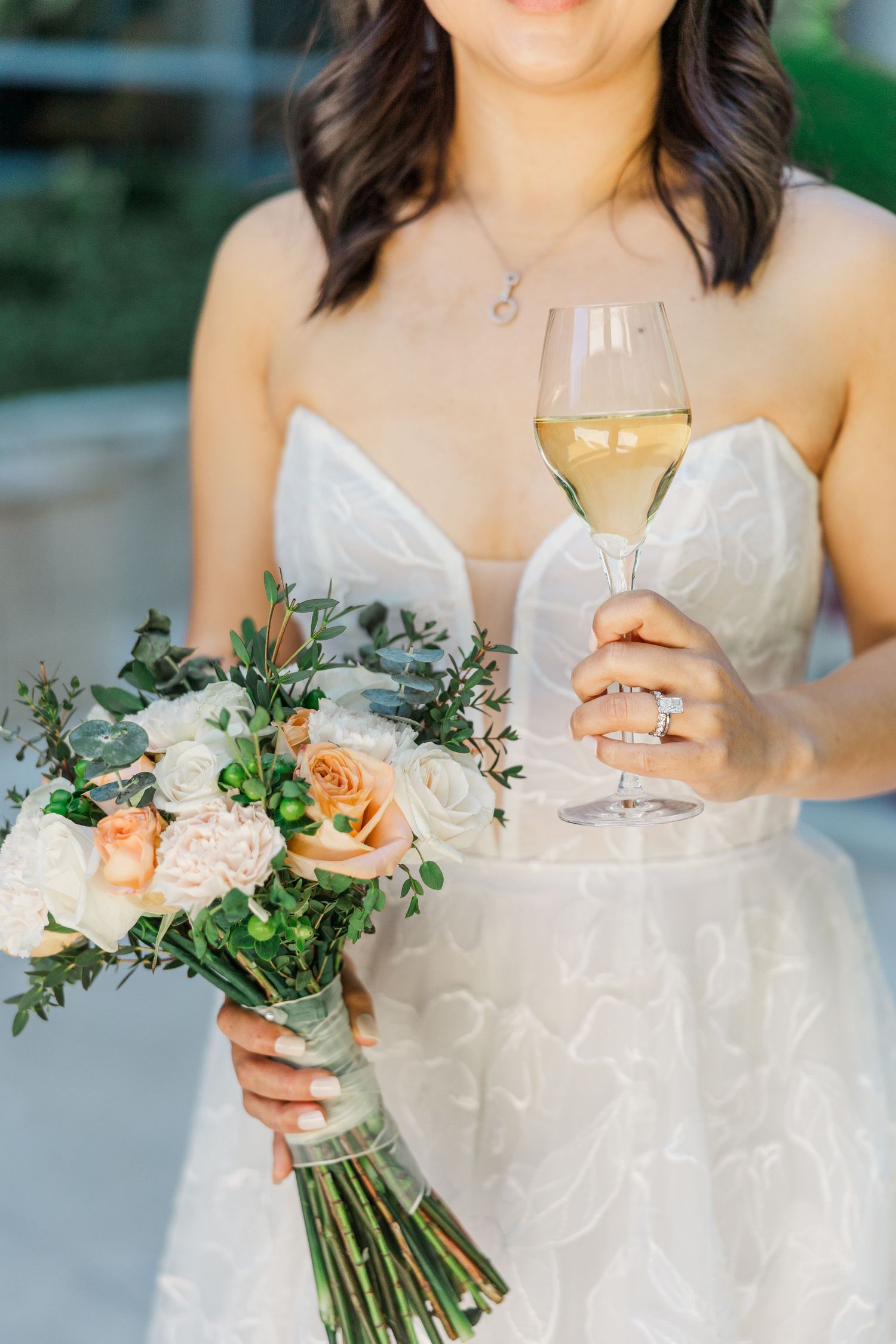 Bride in white gown holding peach and white rose bouquet with greenery and a glass of white wine.