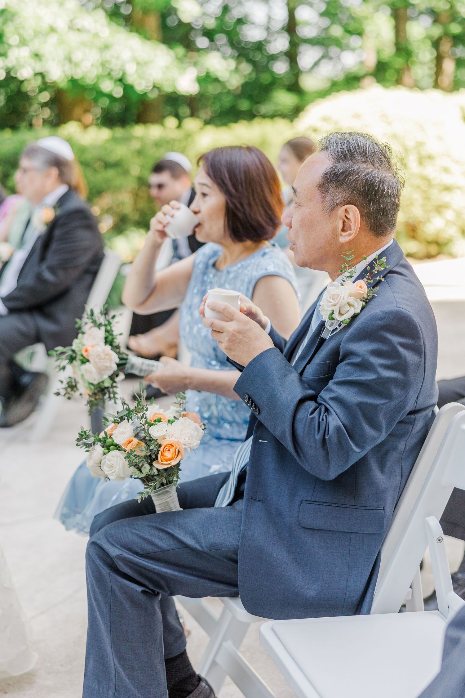 Wedding guests in formal attire sip drinks while seated at an outdoor ceremony surrounded by lush greenery.