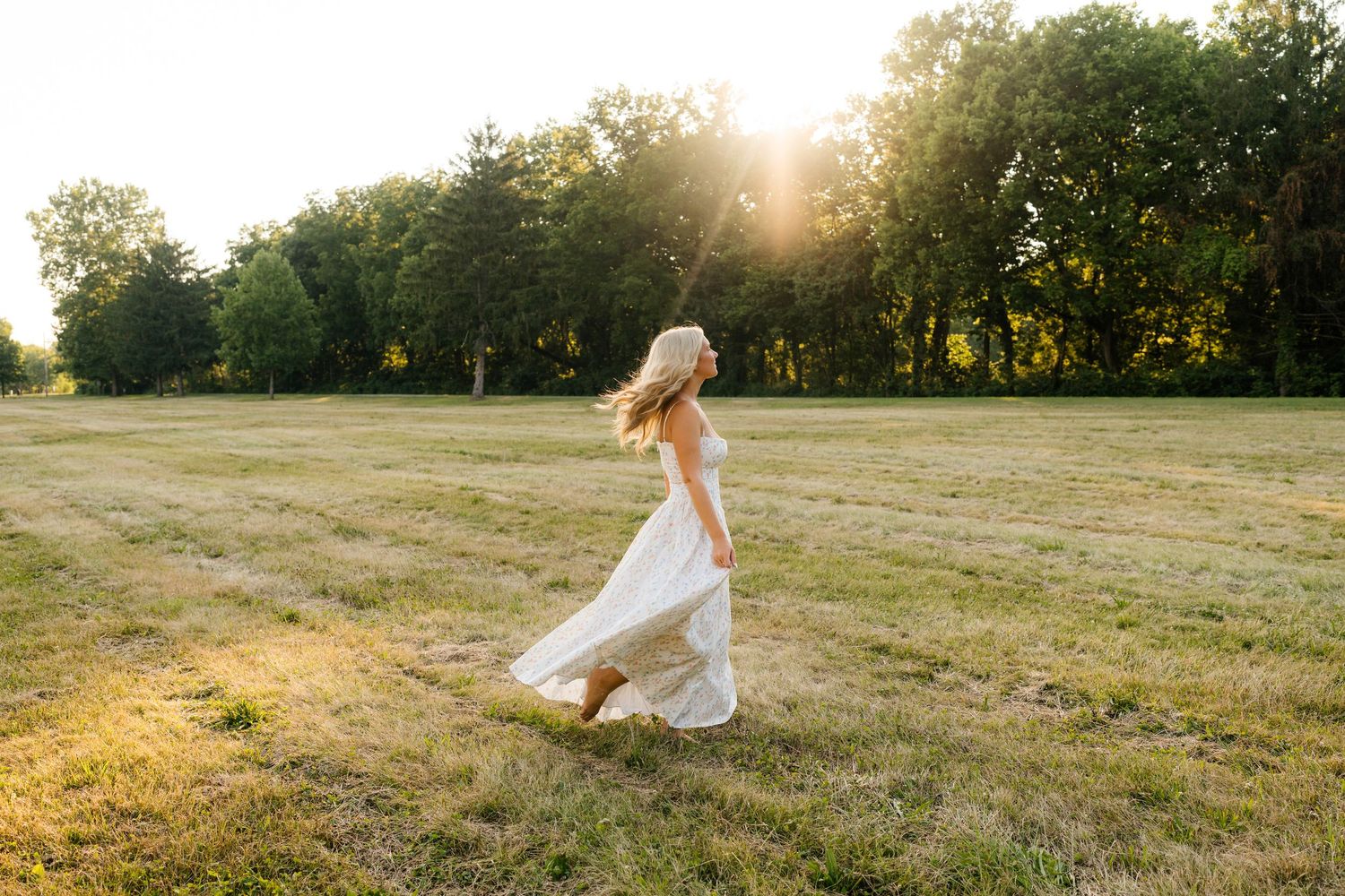 A blonde woman in a flowing white dress walks through a sunlit green field with trees in the background.