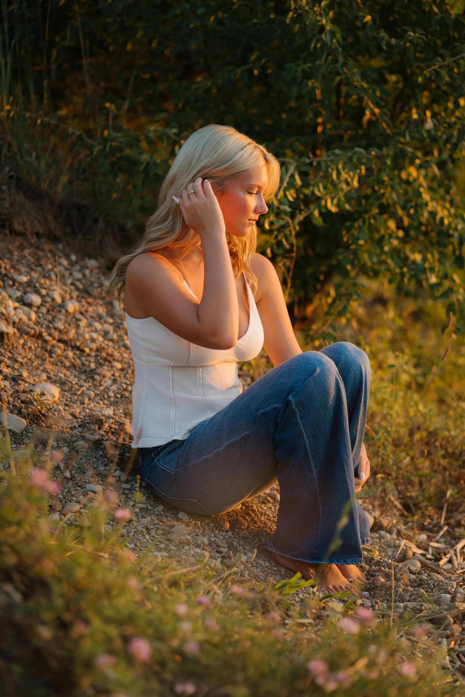 Blonde woman in white tank top and flare jeans sits barefoot outdoors in golden hour sunlight near green bushes.