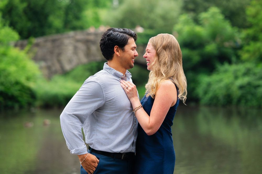 Couple smiling at each other near a scenic pond with a stone bridge and lush greenery in the background.