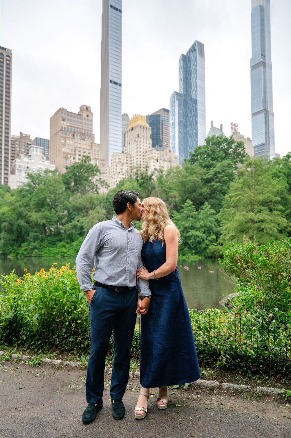Couple sharing a kiss in Central Park with NYC skyscrapers and lush greenery in the background.