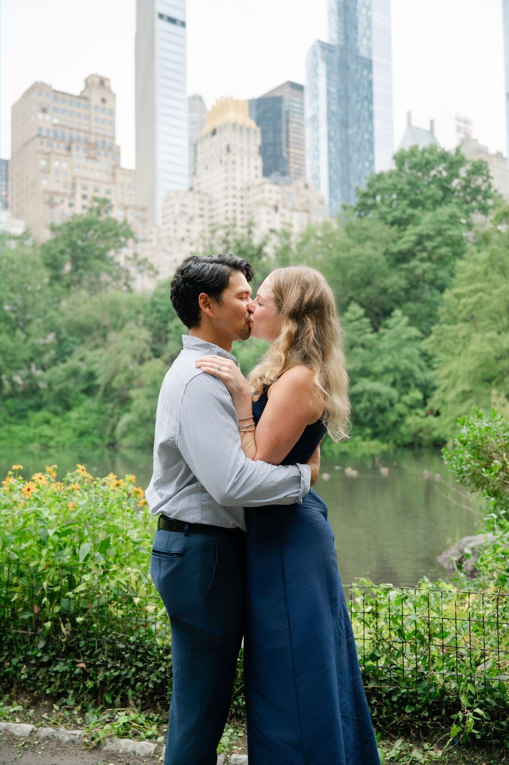 Couple sharing a romantic kiss in Central Park with lush greenery and NYC skyline skyscrapers in the background.