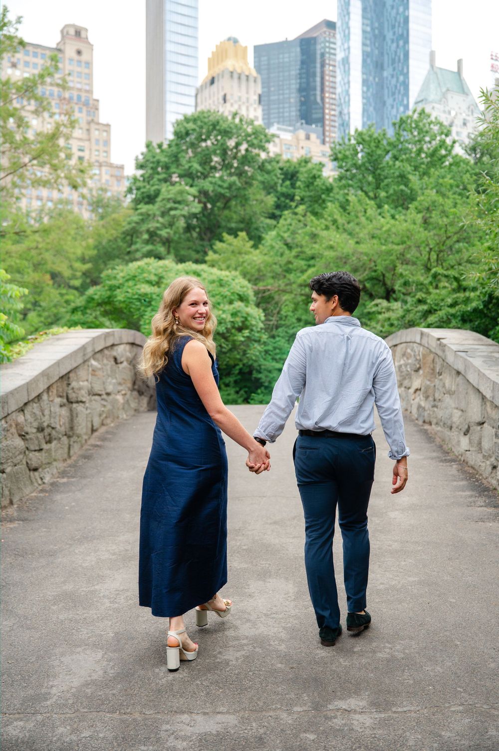 Couple holding hands walking on a stone bridge in Central Park, NYC, with city skyline and green trees in background.
