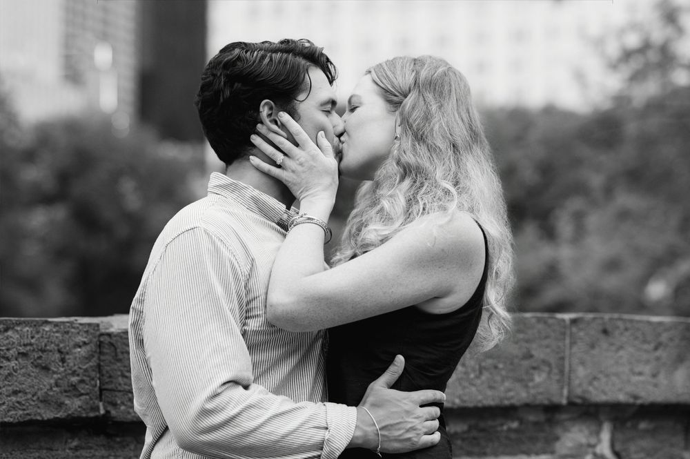Black and white photo of a couple sharing a romantic kiss outdoors with a city skyline in the background.