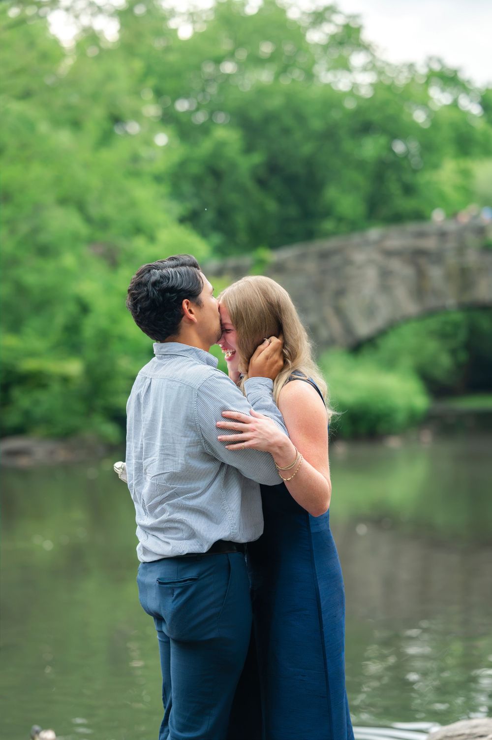 Couple sharing a romantic kiss near a serene pond with a stone bridge and lush green trees in the background.