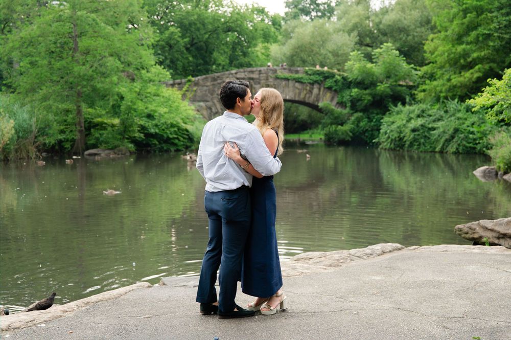Couple kissing by a serene pond with a stone bridge and lush green trees in Central Park, New York.