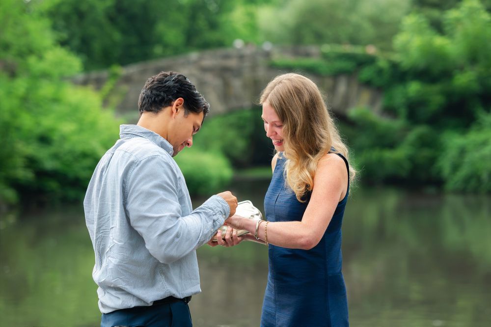 Man placing engagement ring on woman's finger by a scenic pond with stone bridge and lush greenery in background.