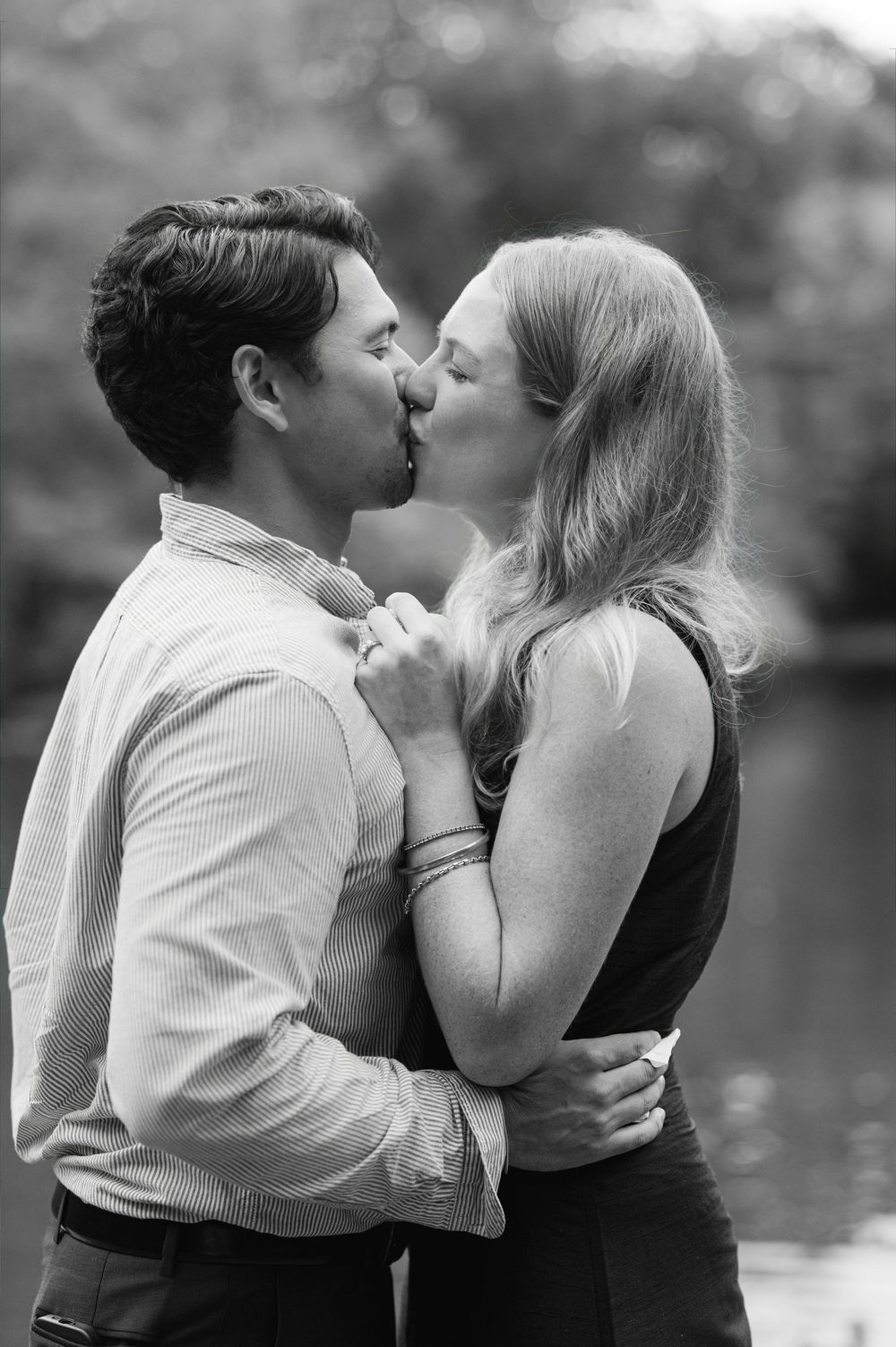 Black and white photo of a couple sharing a romantic kiss outdoors near a serene water backdrop.