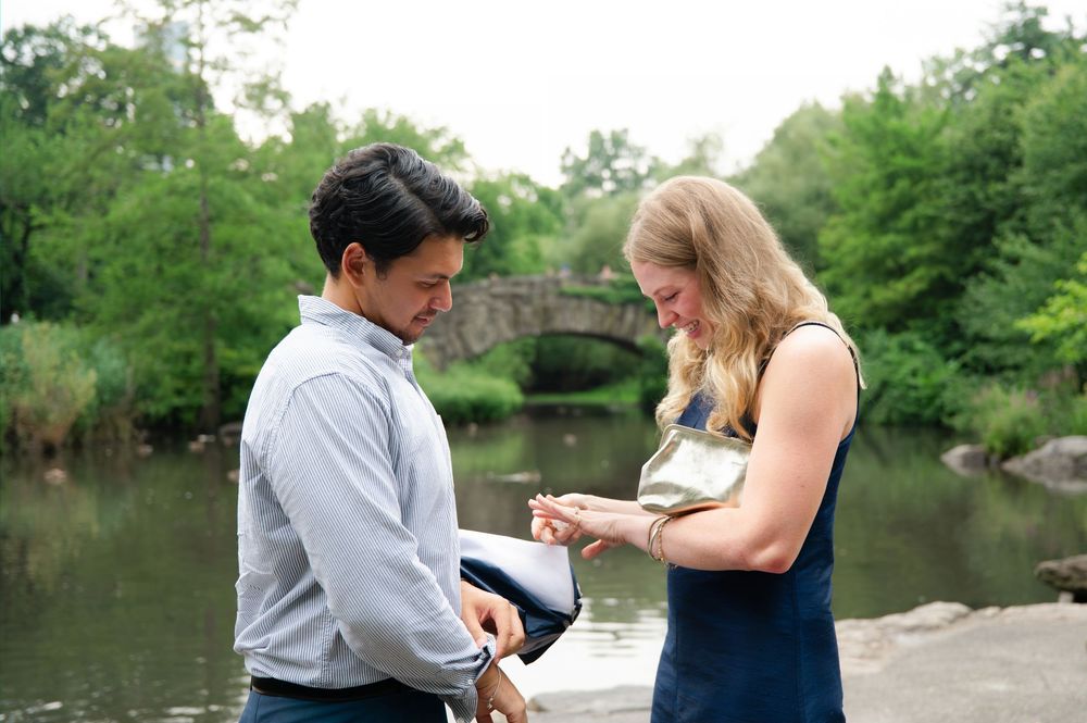 Couple sharing a romantic marriage proposal moment by a pond in Central Park, with a stone bridge and trees in background.