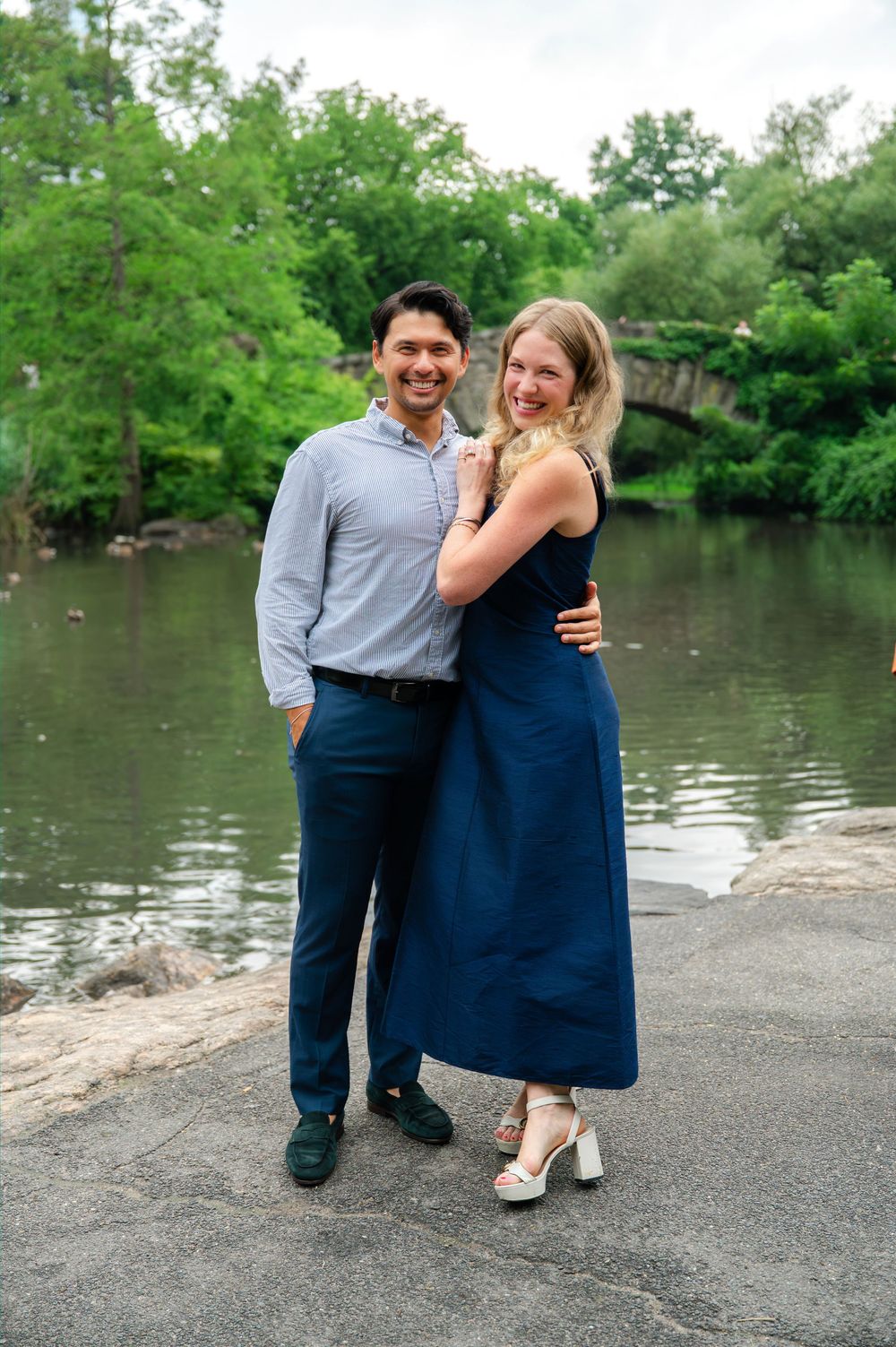Couple posing by a lake in Central Park, woman in navy dress and man in blue shirt, smiling together outdoors.