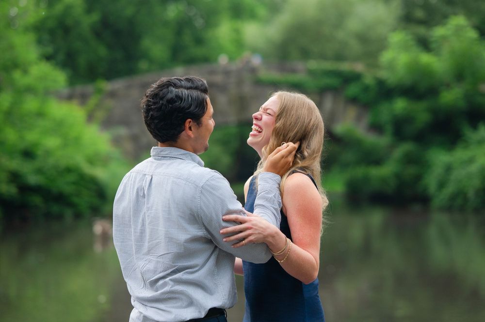 Happy couple laughing together near a stone bridge and pond in a lush green park setting.