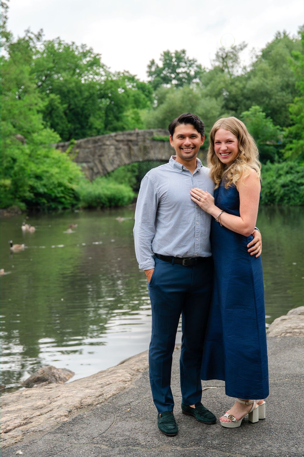 Couple posing and smiling by a pond in Central Park, man in blue pants and woman in navy dress.