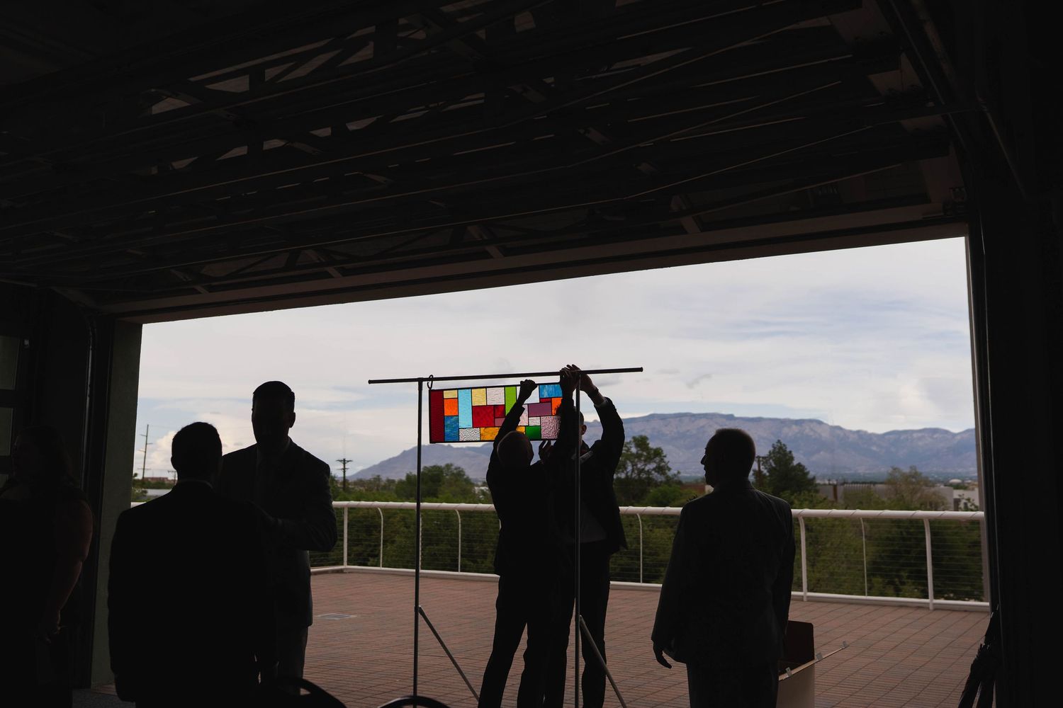 Silhouettes of people holding colorful stained glass panel against mountain backdrop in open warehouse space.