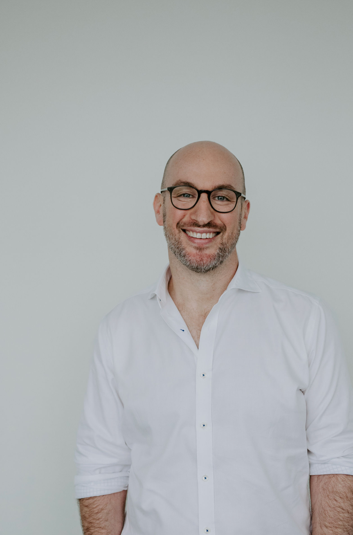 Smiling bald man with glasses wearing a white button-up shirt poses against a light gray background.