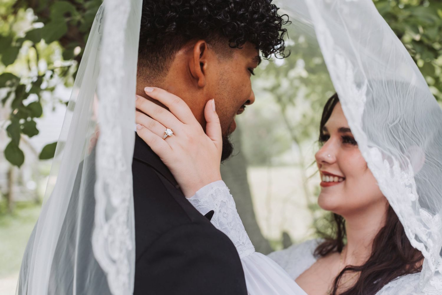 A smiling bride and groom share an intimate moment under a white lace veil outdoors on their wedding day.