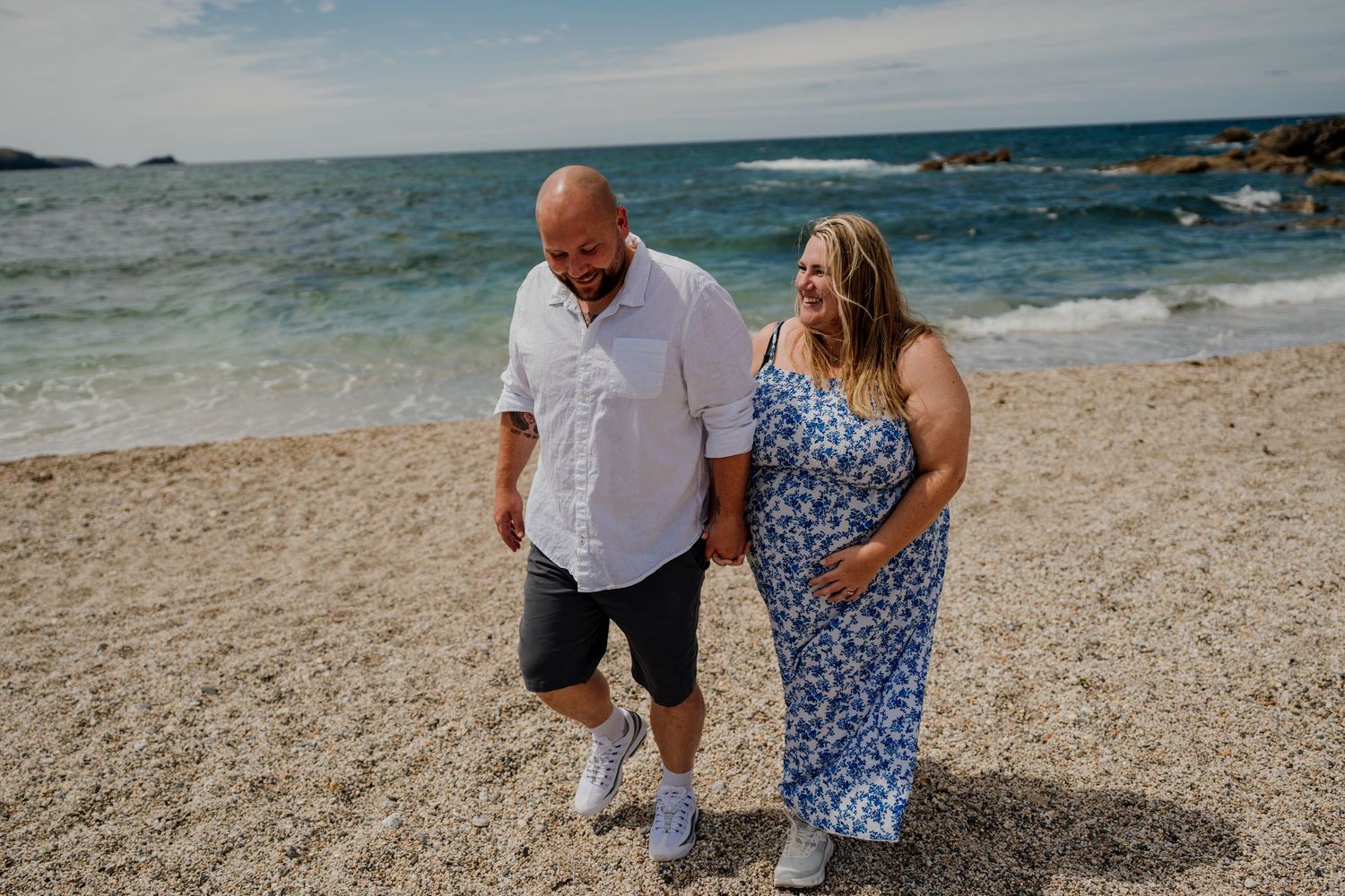 A couple walks hand-in-hand on a sandy beach, smiling, with ocean waves and rocks in the background.