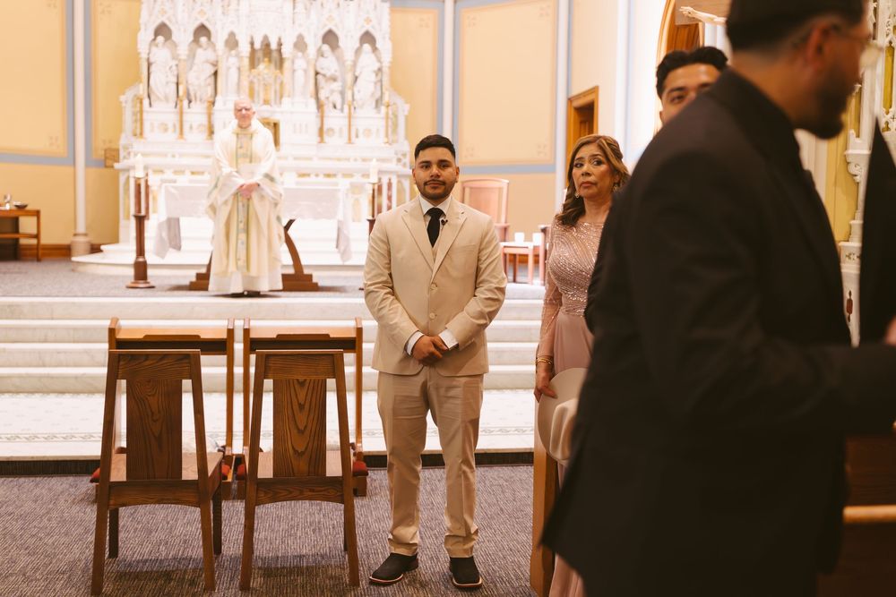 Groom in beige suit stands at church altar awaiting bride during wedding ceremony.