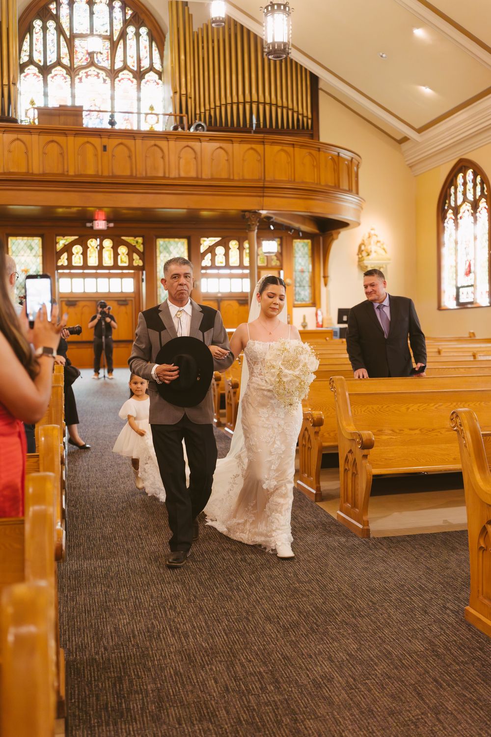 Bride walks down church aisle with escort and flower girl during wedding ceremony.