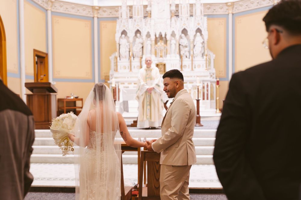 Bride and groom exchange vows at ornate church altar, bride in white gown with veil holding bouquet.